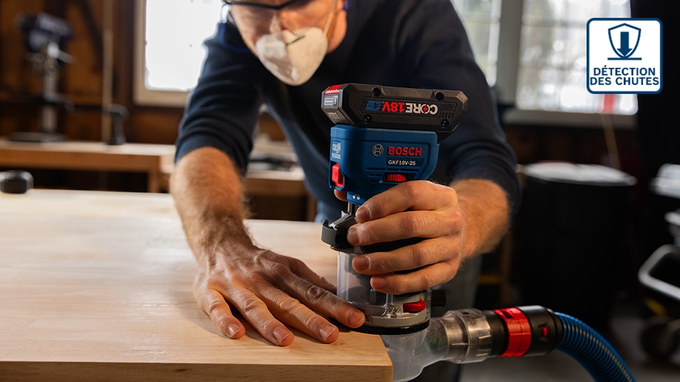 Un homme en chemise bleue et masque utilise une défonceuse Bosch Professional sur un établi en bois, avec des outils en arrière-plan.