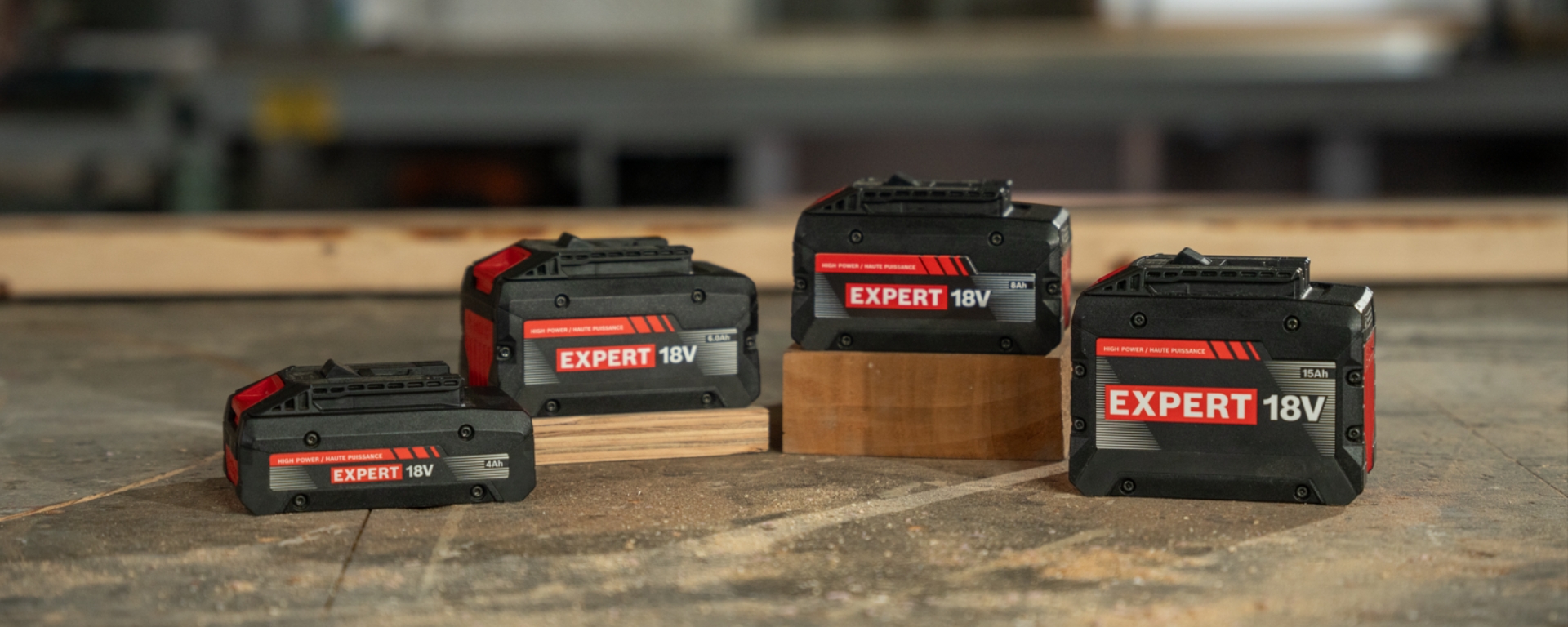 Four Bosch Expert 18V batteries are arranged on a wooden surface, two on blocks of wood, featuring a black and red design.