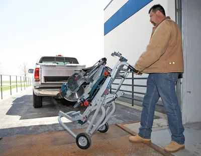 Man wearing a brown jacket and jeans pushing a Bosch saw on wheels towards a truck.