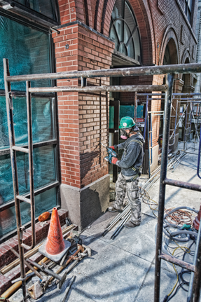 Person in green helmet, gray jacket, and camo pants operating a drill on a brick wall, safety cone and tools visible on the ground.