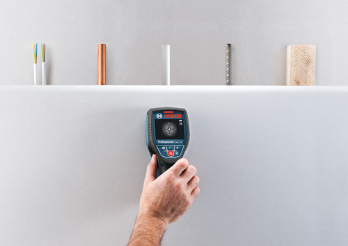 Bosch Professional D-tect 120 detector held by a person, detecting objects behind a wall, with electrical wiring, pipe, metal rod and wood sample in the background.
