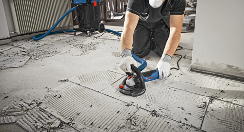 Person wearing gloves and a mask using a Bosch CSG 15 grinder on floor tiles; a vacuum cleaner and debris are also visible.