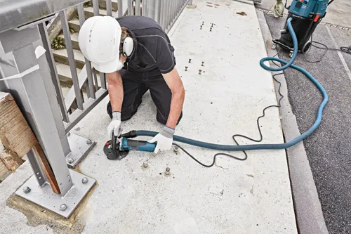 Person wearing safety gear using a Bosch CSG 15 grinder with a hose connected to a vacuum cleaner.