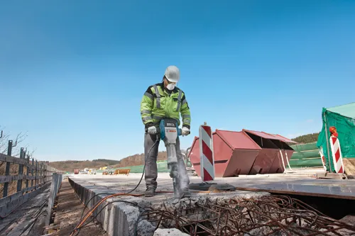 A person wearing a hard hat, safety glasses, and face mask is using a Bosch Brute Turbo breaker to break concrete, with containers, a traffic barrier, and a fence in the background.