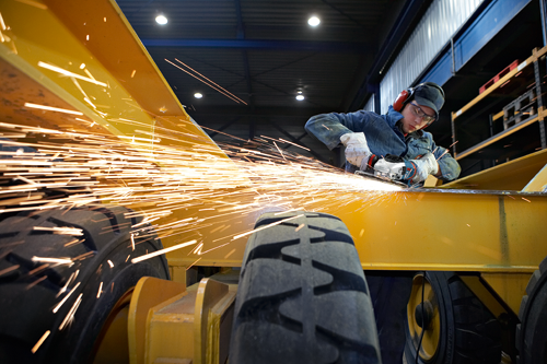 Person using a power tool to cut a metal beam, with sparks flying. A partially visible tire and rim are in the foreground.