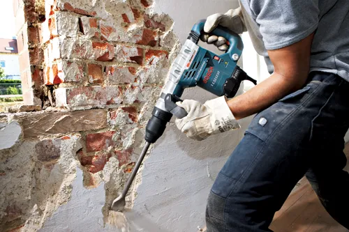 A person using Bosch 11321EVS hammer drill to chip away at brick wall and plaster.
