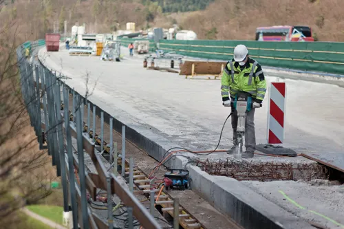 A construction worker in a white helmet and reflective jacket uses a Bosch BRUTE TURBO hammer drill on a concrete surface; a red and white striped sign stands nearby.