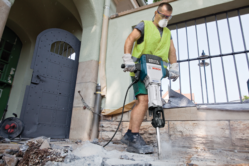 A person wearing protective gear operates a "Brute Turbo" Bosch Boschhammer tool, breaking concrete. The person wears a yellow vest, gloves, and a face mask.