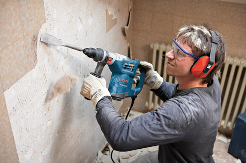 A person wearing safety glasses and ear protection uses a Bosch RH432VCQ demolition hammer to remove tiles from a wall.