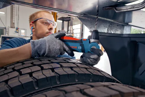 Person wearing safety glasses and earplugs drills into metal using a Bosch power drill while wearing gloves, with tire in foreground.