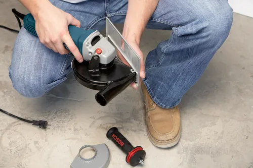 Person wearing blue jeans holding a Bosch angle grinder, with a black and red "BOSCH" handle and a gray guard on the ground.