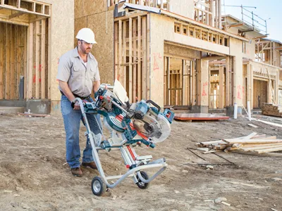 A construction worker, wearing a hard hat, carries a Bosch power tool on a wheeled cart near a building under construction.
