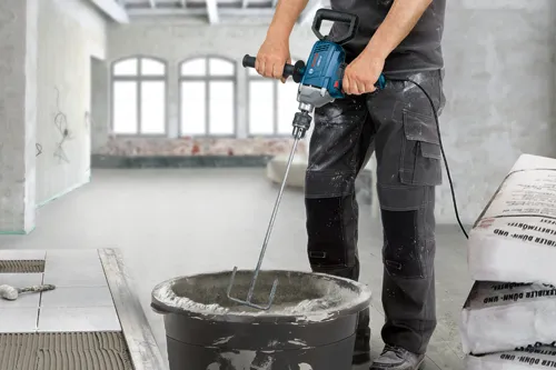 Bosch GBM 16-16 mixer, held by a person, mixing mortar in a bucket. A bag of "FLEXIBLER DÜNN- UND..." is visible. Tiles and tools are on the floor.