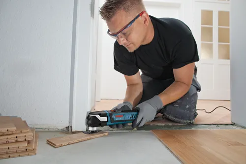 Man wearing safety glasses, gray gloves, and a black shirt kneels, using a Bosch GOP40-30 to cut a wooden board.