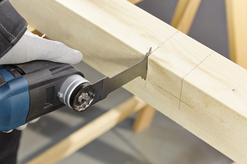 Person wearing gray work gloves using a saw to cut into a wooden beam.