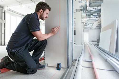 Man kneeling, drawing on drywall near Bosch laser level.