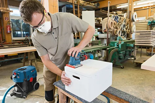 A person wearing safety glasses and a mask is sanding a white box with a Bosch GSS 20-40 sander. A Bosch vacuum cleaner is visible.