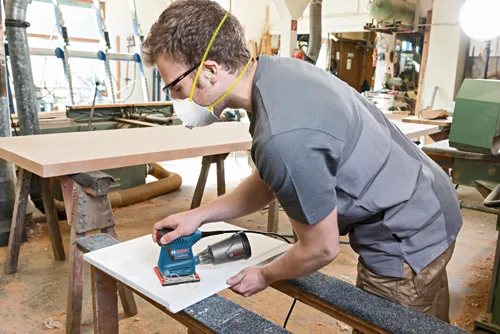 Man sanding a white board with Bosch GSS20-40 sander, wearing safety glasses, ear plugs, and a respirator mask.