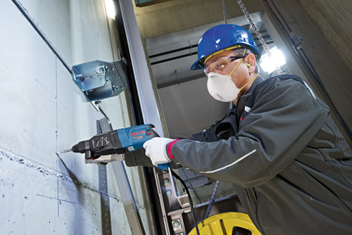 A person wearing a hard hat, safety glasses, and face mask using a Bosch Bulldog Xtreme hammer drill to drill into a wall.