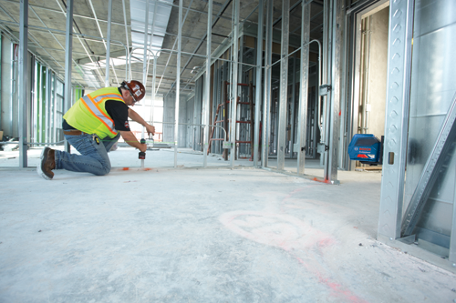 A person wearing safety gear, kneeling on a concrete floor, using a laser level to work on metal framing. The Bosch Professional level is attached to the metal frame.