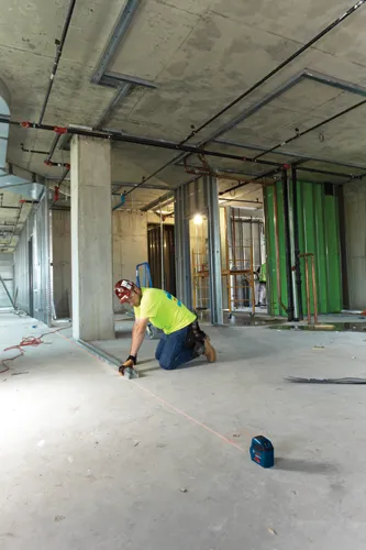 Person kneeling on concrete floor using laser level, with metal studs, columns, and piping overhead; Bosch Professional laser level on the floor.