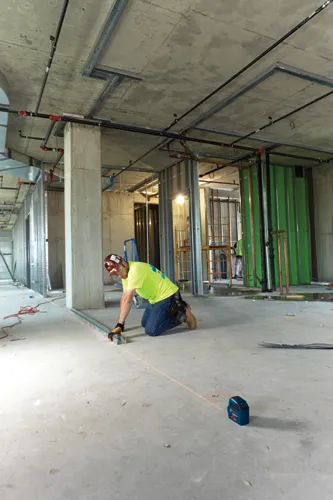 Construction worker kneeling, installing metal frame along a red laser line. Bosch laser level on the floor, concrete ceiling with pipes.