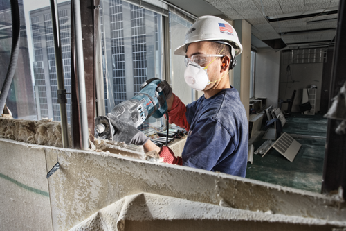 A person wearing safety glasses, a respirator, and a hard hat with an American flag is using a reciprocating saw to cut into a wall.