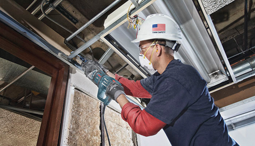 A worker, wearing a hard hat with a USA flag and the text "THINK SAFETY", safety glasses, and a mask, uses a reciprocating saw.