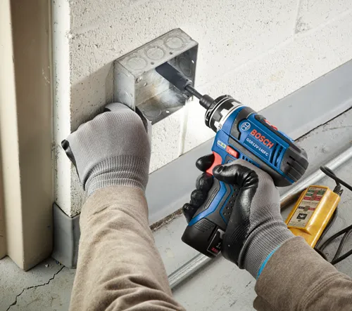 Bosch GSR12V-140FC cordless drill being used to drill into a metal electrical box, with a voltage tester visible.