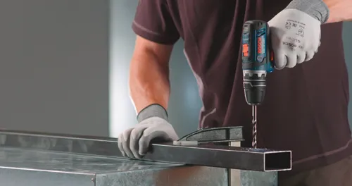 A person wearing gloves uses a drill to make a hole in a metal piece; the drill is a Bosch GSR12V-140FC.