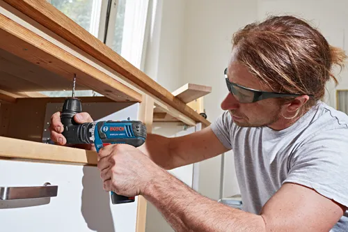 Man wearing safety glasses using a Bosch GSR12V-140FC drill to drill into a wooden shelf.