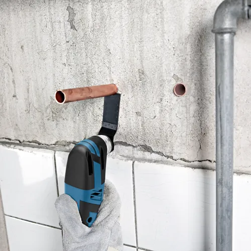 Bosch oscillating multi-tool cutting copper pipe in a wall. A hand in a work glove holds the tool.
