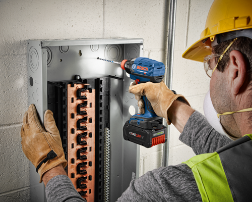 A person wearing gloves, safety glasses, a mask, and a yellow hard hat drills into an electrical panel with a Bosch Lithium-Ion 18V drill.