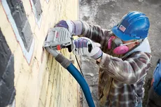 Person using a Bosch grinder on a brick wall, wearing a hard hat with US flag sticker, respirator, gloves, and plaid shirt.