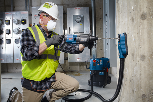 A person wearing safety gear is drilling a hole in a concrete wall using a blue and black Bosch BULLDOG X-Flex rotary hammer and dust collection attachment; a HEPA Ready vacuum is connected.