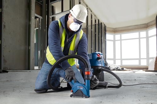 Person in safety gear kneels, using a Bosch GASODC 400 concrete saw attached to a vacuum in a construction site.