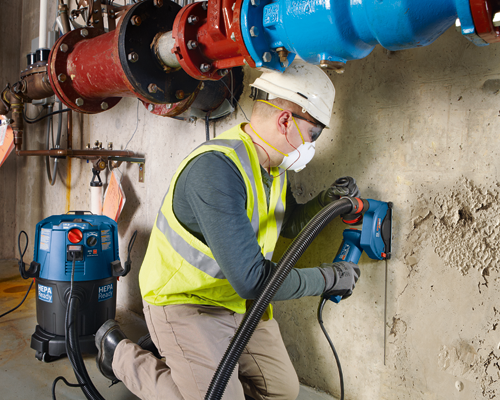 Man wearing safety gear using a Bosch tool on a concrete wall, a HEPA Ready vacuum is nearby.