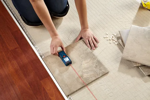 Person using Bosch BLAZE laser measure to measure a tile, on a tile-laying surface, with other tiles nearby.