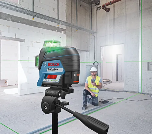 Bosch Professional GLL3-330CG laser level on a tripod, projecting green lines, with a person wearing a safety vest and hard hat using a smartphone in a construction site.