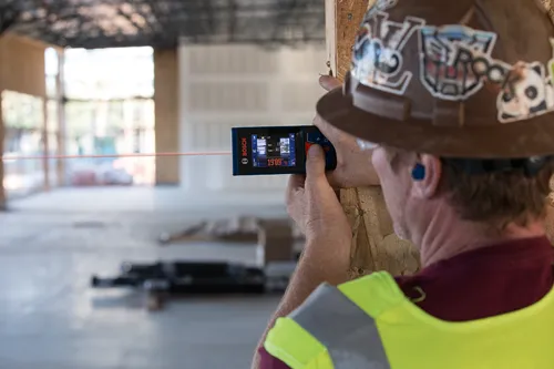 A person wearing a hard hat, safety vest, and earplugs is using a Bosch distance measuring tool. The tool displays "19’09” ½”.