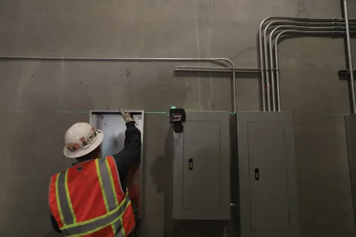 A construction worker in an orange safety vest and hard hat uses a level to align equipment on a wall. A Bosch laser level is visible.