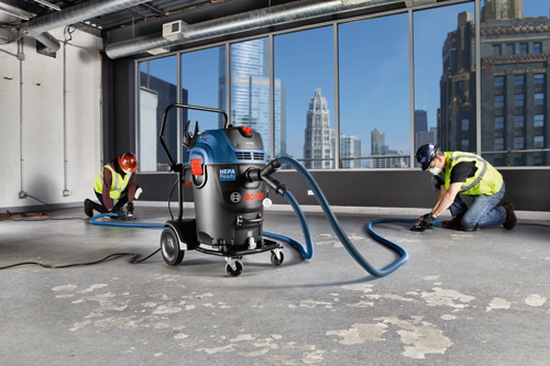 Two construction workers in safety gear are using a Bosch HEPA Ready vacuum, with a city skyline in the background.