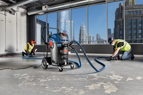 People using power tools connected to a Bosch HEPA Ready vacuum in an unfinished space with city views.