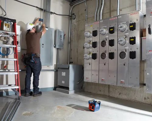 Person wearing a white hard hat working on electrical panel, with "WARNING 480 VOLTS" labels, with a red ladder and a Bosch laser level on the floor.