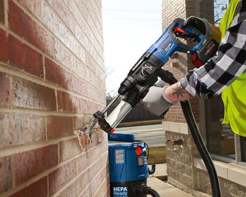 Bosch BULLDOG GBH18V-26D cordless rotary hammer drill being used to drill into a brick wall. A second tool, a blue Bosch HEPA Ready vacuum, is connected to the hammer drill. A person wearing a checkered shirt, safety vest, and work gloves holds the drill.