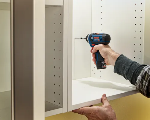Hand holding a Bosch cordless drill, attaching a screw to a white cabinet. "BOSCH LITHIUM-ION 12 2.0 Ah Li" is visible on the drill.