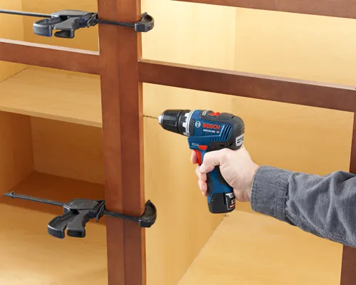 Bosch GSR12V-300 cordless drill being used to drill into a cabinet with clamps attached.