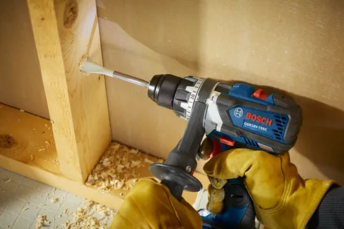 Bosch GSR18V-755C drill drilling into wood, held by gloved hands.
