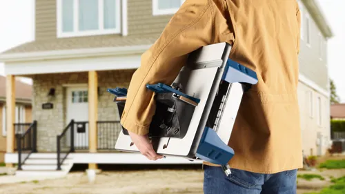 A person wearing a tan jacket and jeans carries a power tool with blue and black accents near a house.