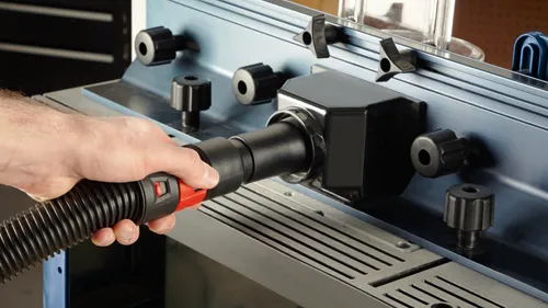 Hand holding a black and red vacuum hose connected to a black rectangular device with knobs, on top of a metal surface.